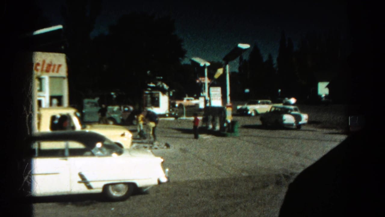1967 Colorado home-movie frame showing a dimly lit gas station, classic cars, and people near the pumps at night.