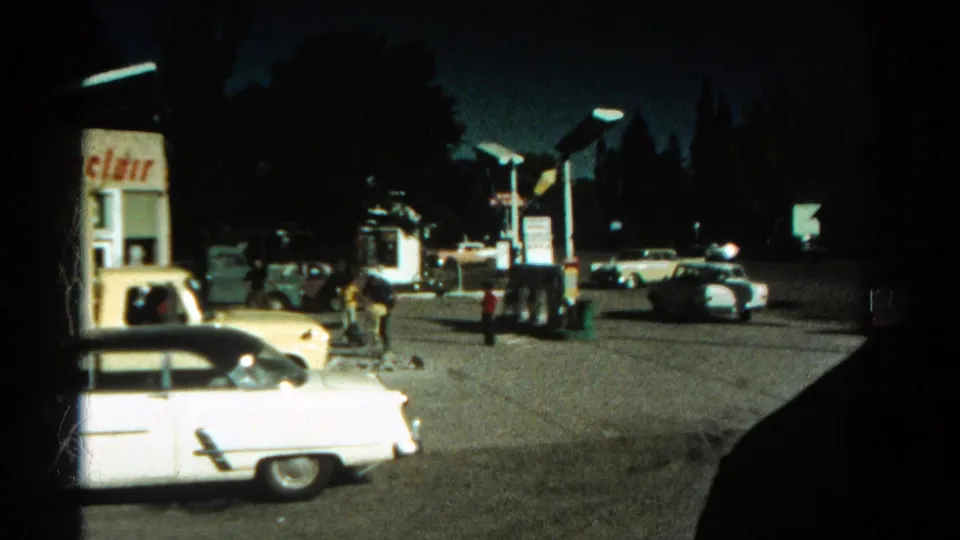 1967 Colorado home-movie frame showing a dimly lit gas station, classic cars, and people near the pumps at night.