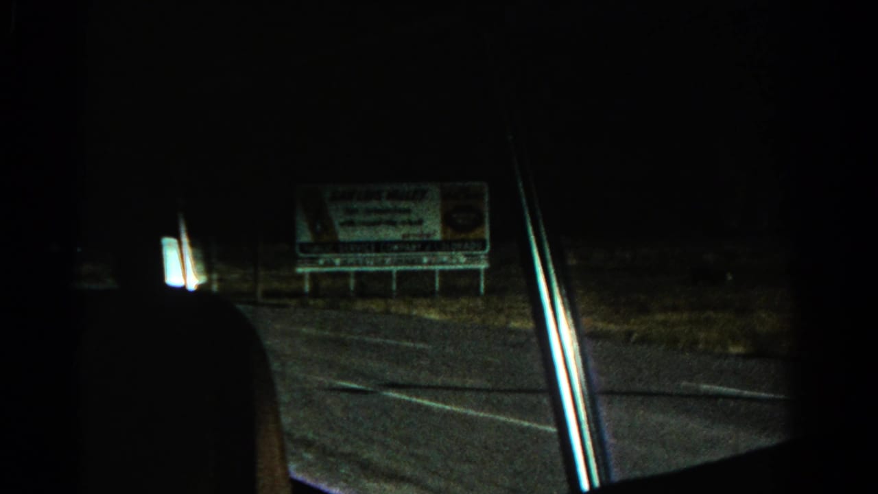 Dark roadside view from inside a vehicle with a billboard and road edge ahead.