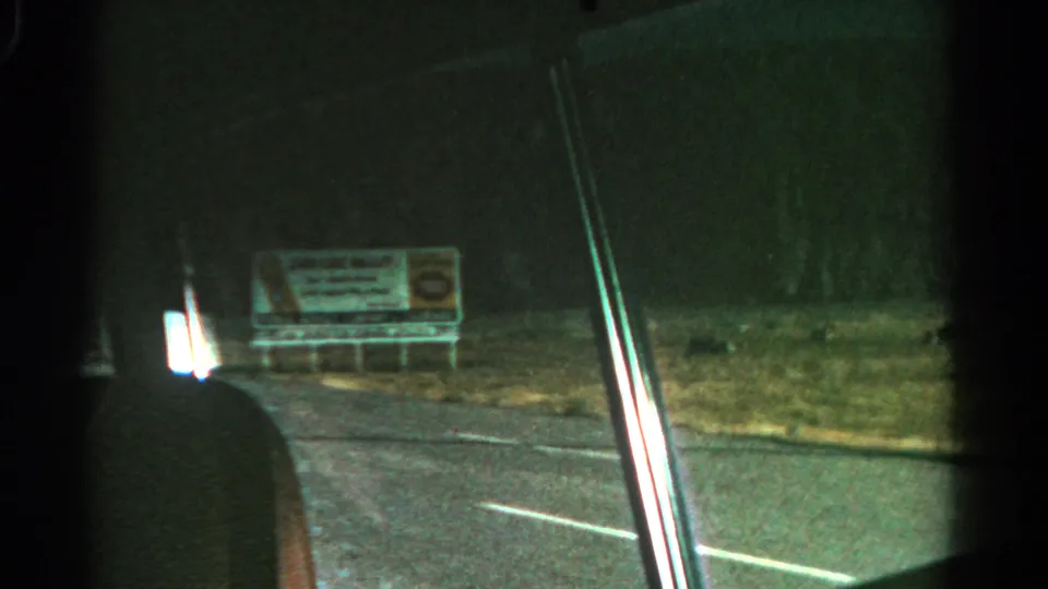 Low-light 8mm frame from a moving car, showing a roadside billboard, grass, pavement, and window edge.