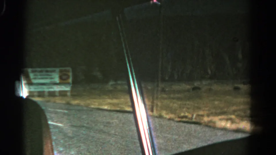 Dark Colorado roadside view from a car, with a billboard, grass shoulder, pavement, and window frame in the foreground.