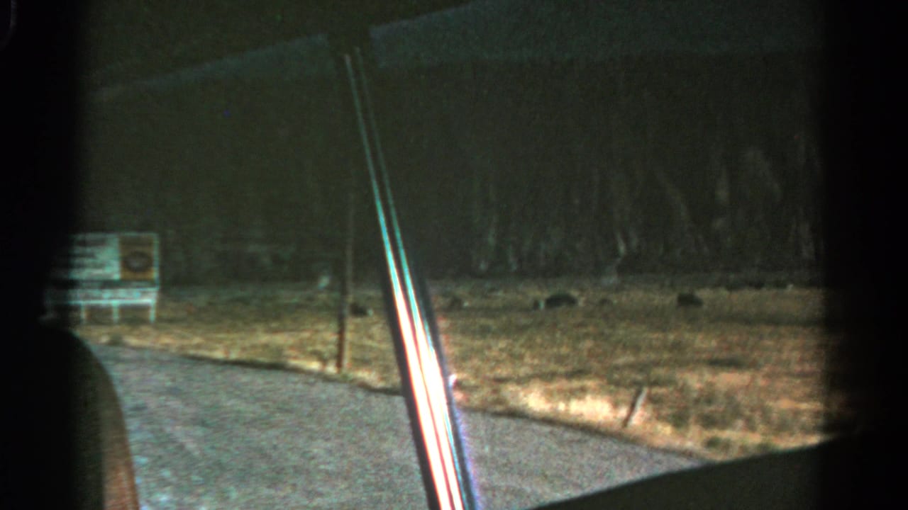 1967 Colorado 8mm frame looking from a moving car past the window edge toward a gravel road, dry field, low hills, and a small sign.