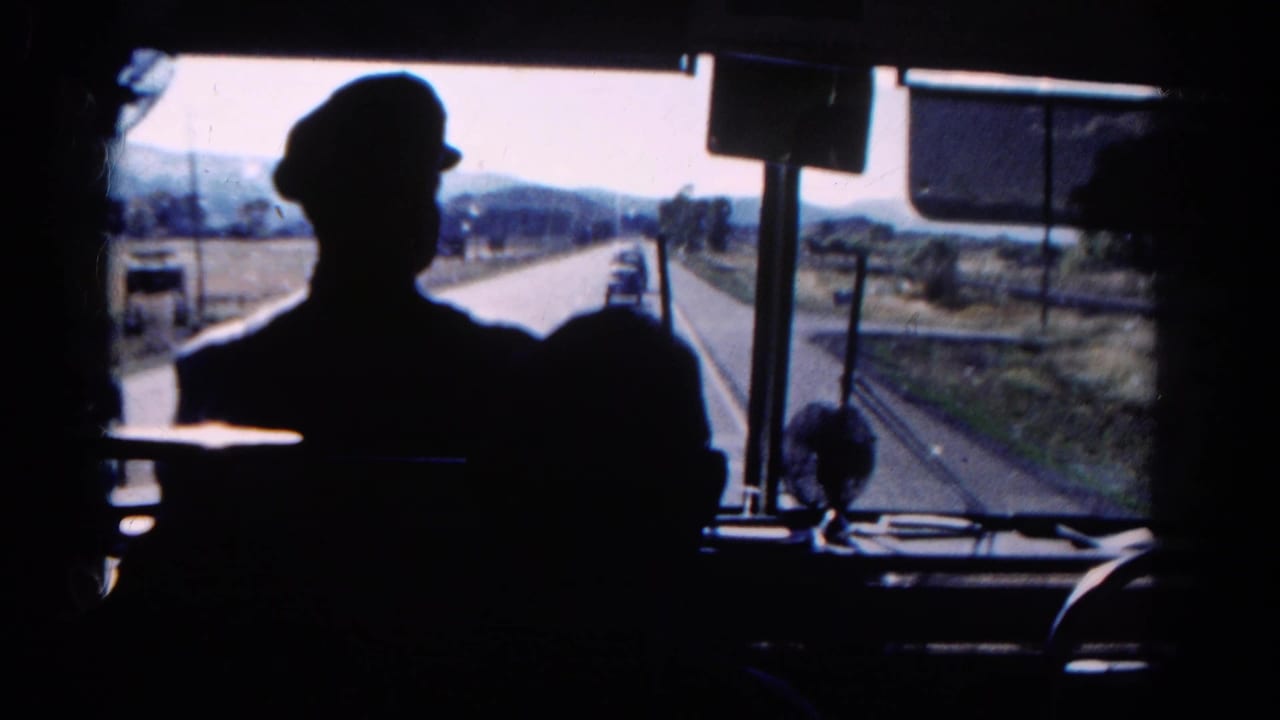 Interior bus view in Colorado, 1967, with a capped driver in silhouette and vehicles visible ahead on a rural highway.