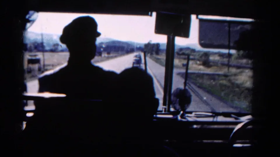 Interior bus view in Colorado, 1967, with a capped driver in silhouette and vehicles visible ahead on a rural highway.