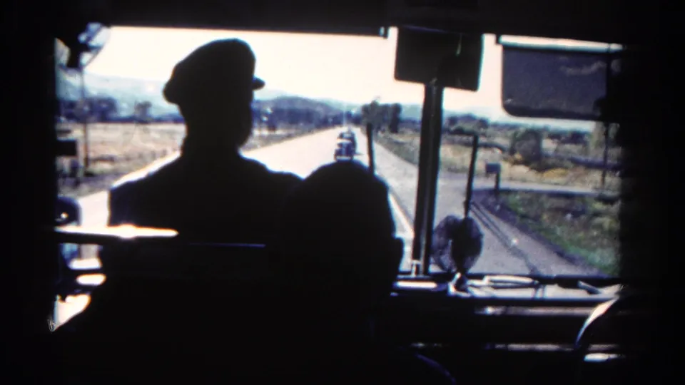 1967 Colorado home-movie frame showing the bus driver's profile silhouette with the highway, fields, and hills beyond the windshield.