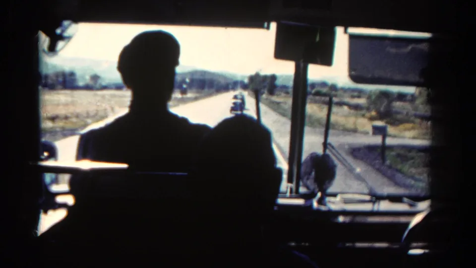 Interior 8mm bus view from Colorado in 1967, looking past the driver's silhouette toward vehicles on a rural two-lane highway.