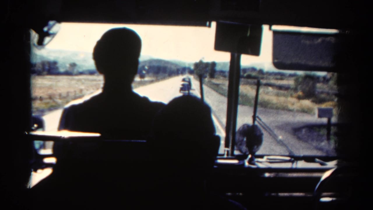 Silhouetted bus passengers seen through a front windshield on a rural road.