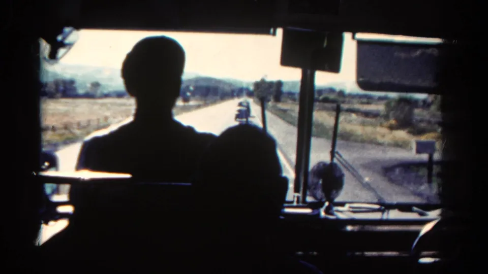1967 Colorado bus interior view with dark passenger silhouettes, a two-lane road, fields, and low hills ahead.