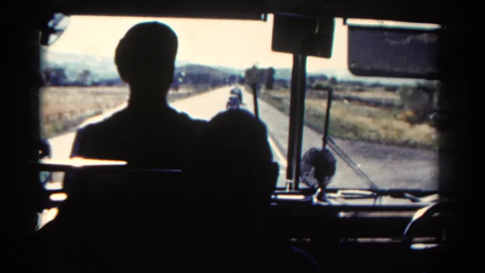 Inside a moving bus in Colorado, silhouetted riders face a rural road bordered by fields and distant hills.