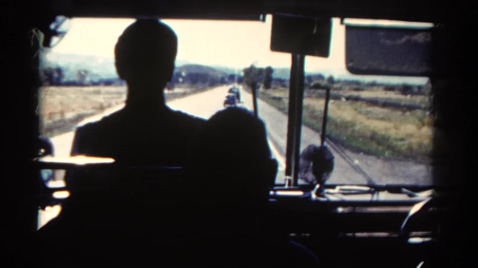 Passenger-level 8mm view from inside a bus, showing silhouetted riders and rural Colorado fields beyond the front glass.