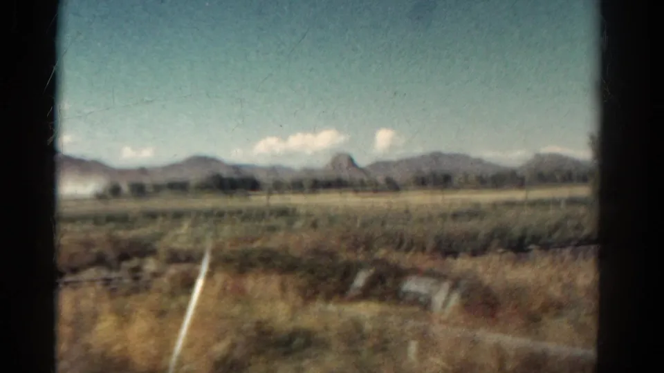 Rural Colorado landscape from a moving vehicle, with open fields, fence lines, distant mountains, and a pale blue sky.