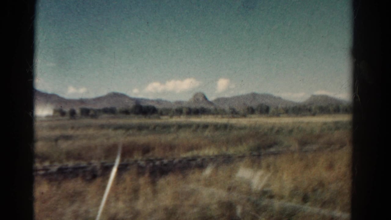 Open field and mountains viewed from inside a moving train, with a white marker near the foreground.