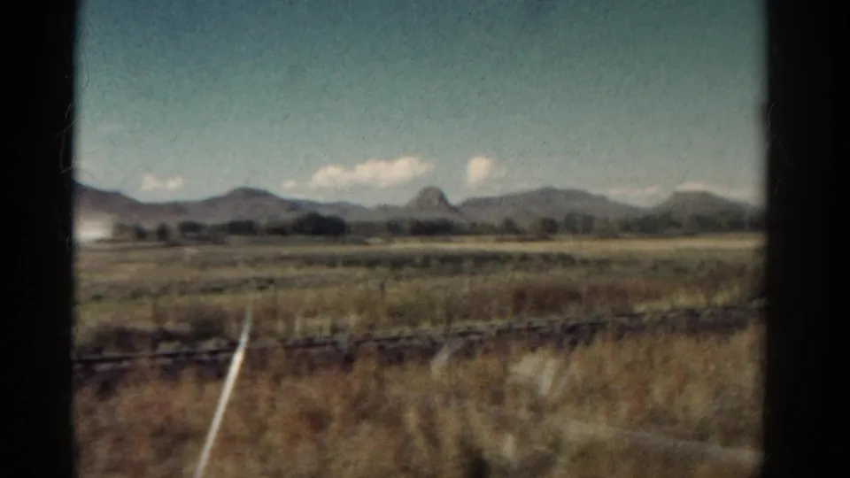 Colorado 1967 home-movie frame from a moving train, showing dry foreground grass, a diagonal white marker, and mountains beyond open fields.