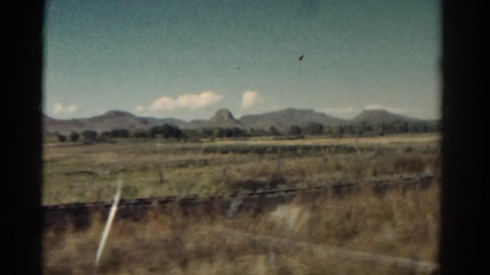 1967 Colorado 8mm view through train glass, with plains, distant mountain silhouettes, pale clouds, and soft reflections across the frame.
