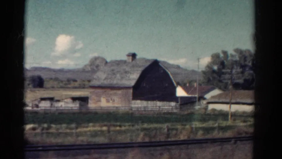 A barn and small rural house pass beside train tracks in 1967 Colorado, with mountain forms and trees behind them.