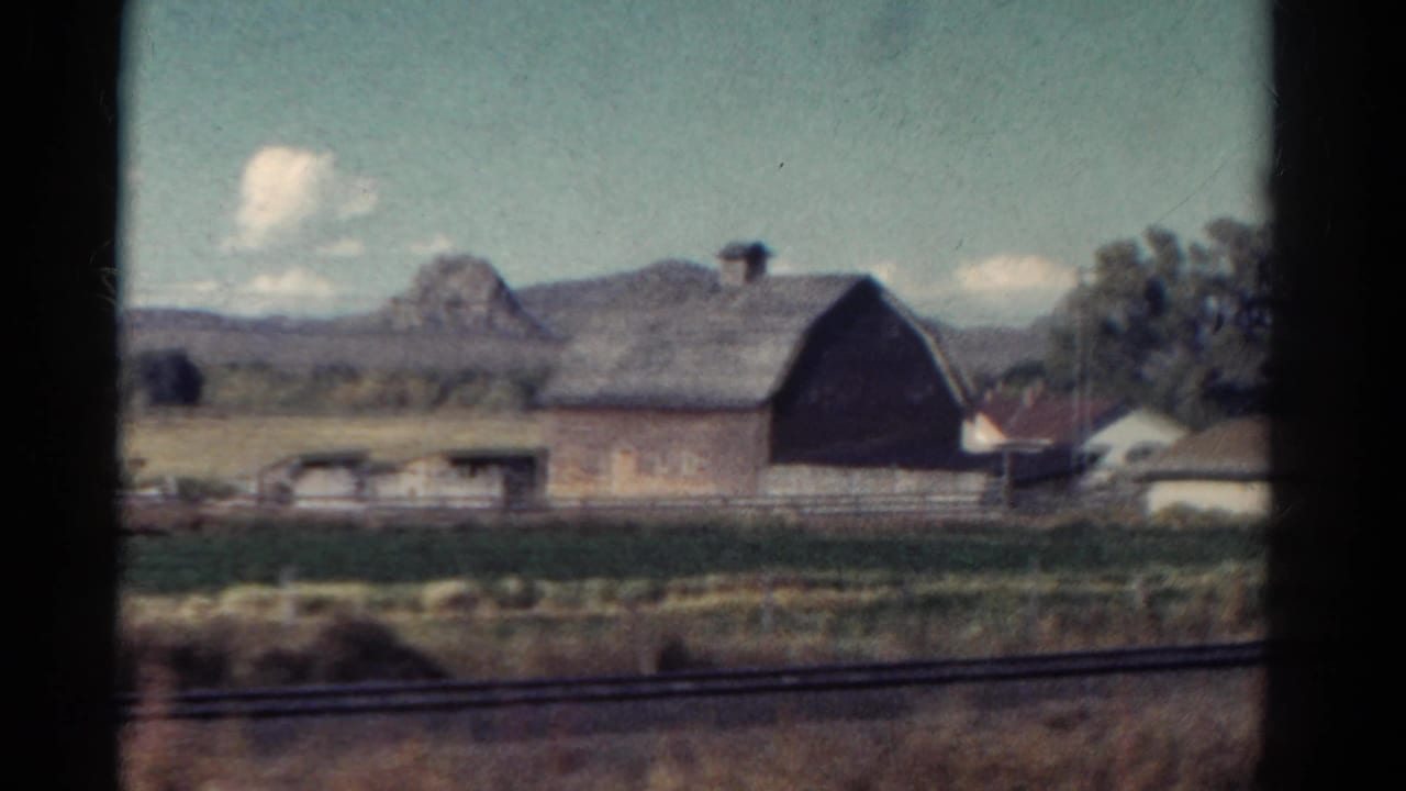 1967 Colorado home-movie frame showing a large barn, fenced farmland, roadside grass, and distant hills from a moving vehicle.