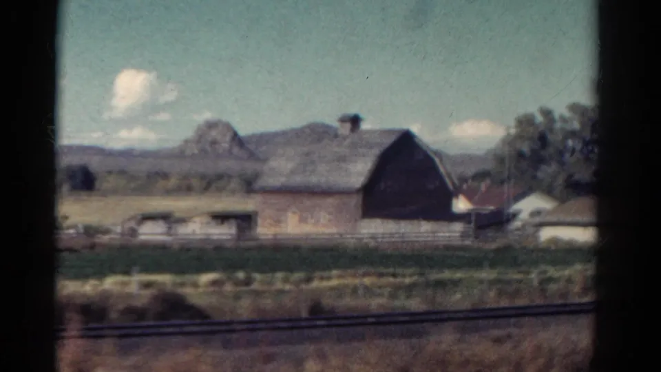1967 Colorado home-movie frame showing a large barn, fenced farmland, roadside grass, and distant hills from a moving vehicle.