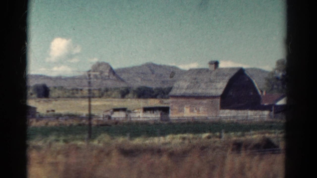 Large barn and farm outbuildings beyond a fence, with fields and low mountains behind.