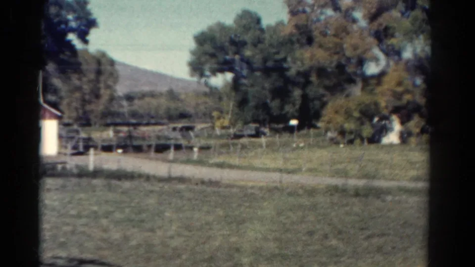 1967 Colorado home-movie frame showing fenced pasture, mature trees, a partial roadside building, and hills beyond.