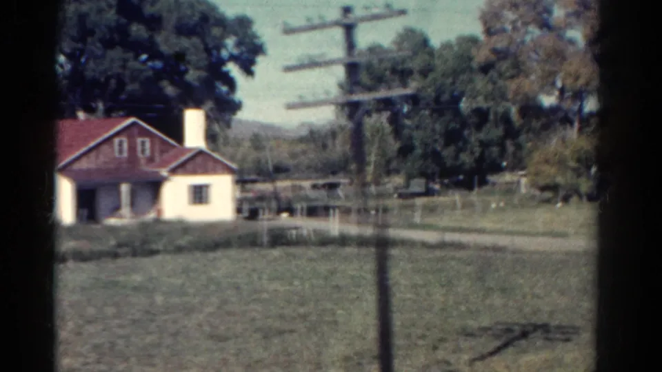 1967 Colorado road-view frame showing a red-roofed farmhouse, fenced fields, utility pole, trees, and low hills.