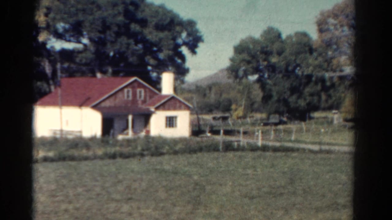 1967 Colorado home-movie frame showing a cream rural house with a red roof, chimney, trees, and field seen from a moving car.