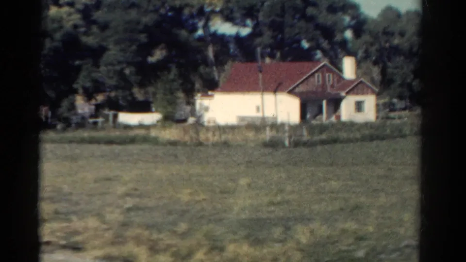 1967 Colorado home-movie view of a cream rural house with a red roof, utility pole, trees, and field during a moving-car pass.