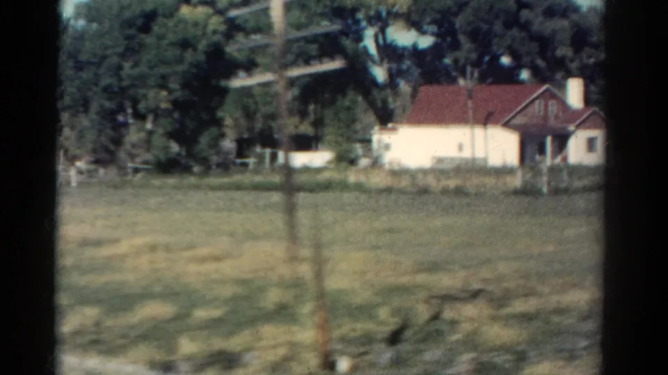 Late frame from 1967 Colorado footage showing the red-roofed house across a field, with foreground motion blur from the moving car.