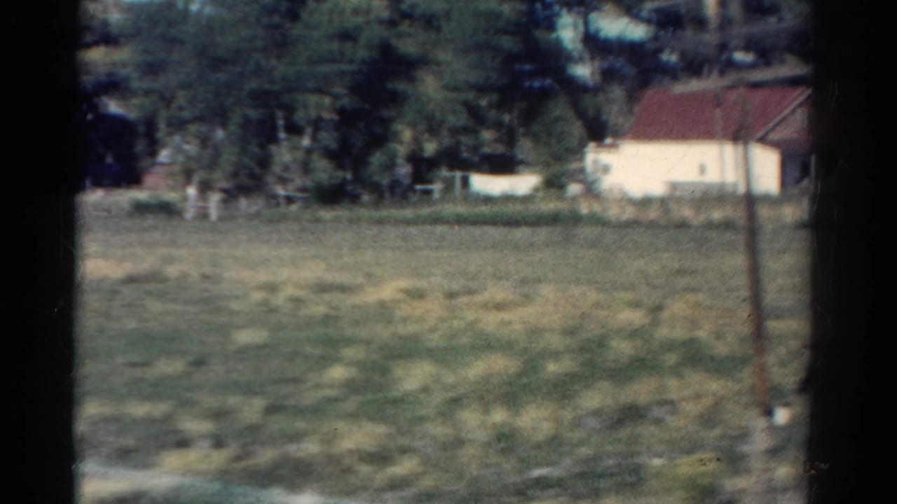 Grassy field with a white rural building, red roof, trees, and film gate edges.