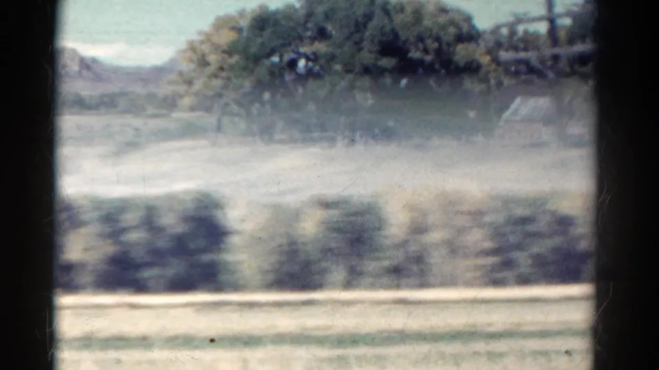 1967 Colorado roadside frame with motion-blurred foreground, trees, distant mountains, and a small rural building at right.
