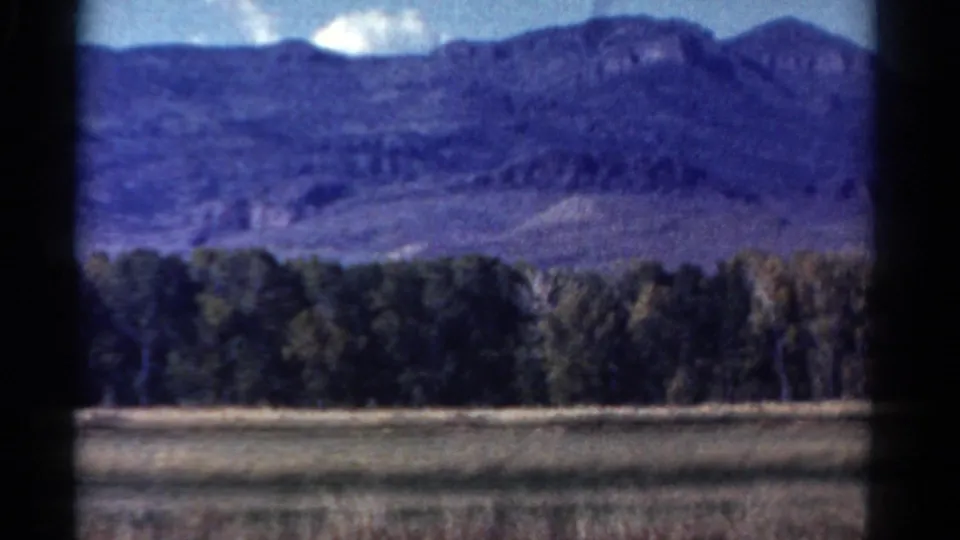 1967 Colorado 8mm frame showing a mountain ridge, blue sky, open field and dense trees from a moving car window.