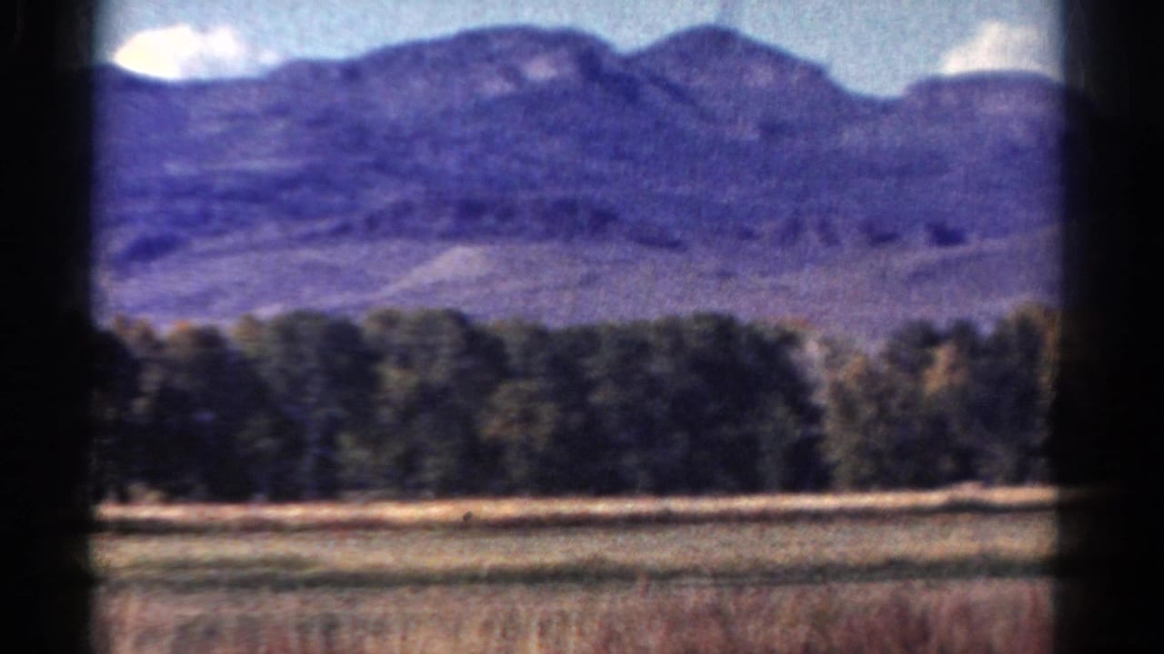 1967 Colorado 8mm frame showing a distant mountain ridge, forest line, dry foreground field, and pale clouds.