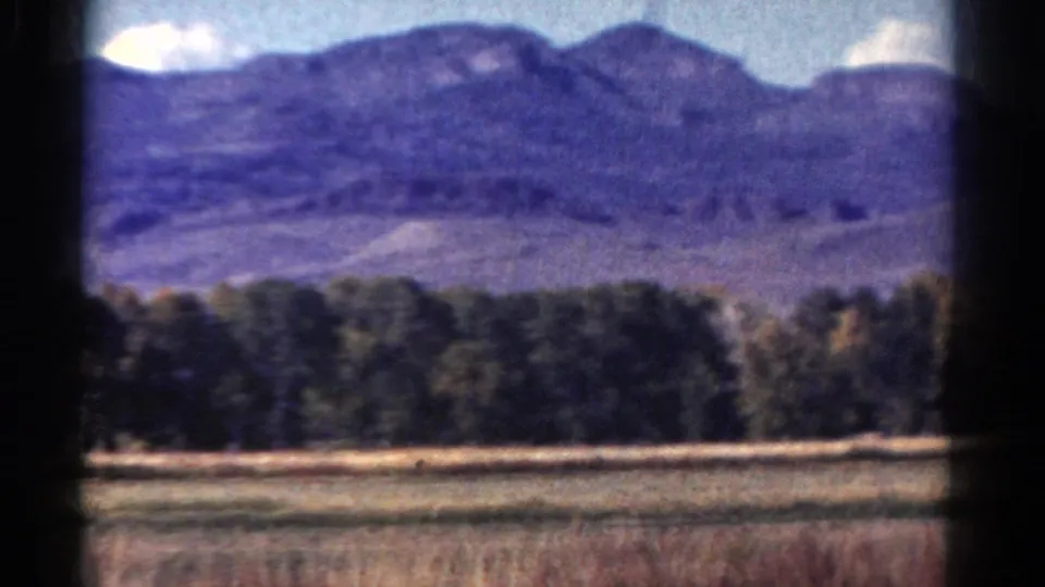 1967 Colorado 8mm frame showing a distant mountain ridge, forest line, dry foreground field, and pale clouds.