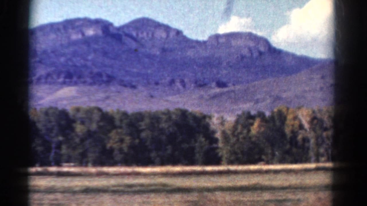 1967 Colorado home-movie view of a broad mountain ridge behind trees and an open field under scattered clouds.