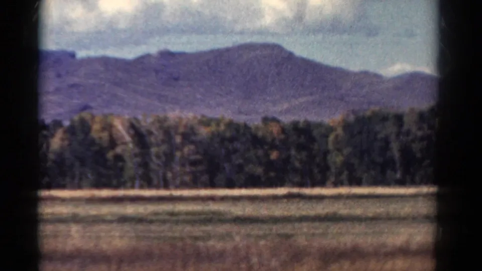 1967 Colorado home-movie frame showing a softened mountain profile beyond trees and a field under cloud cover.