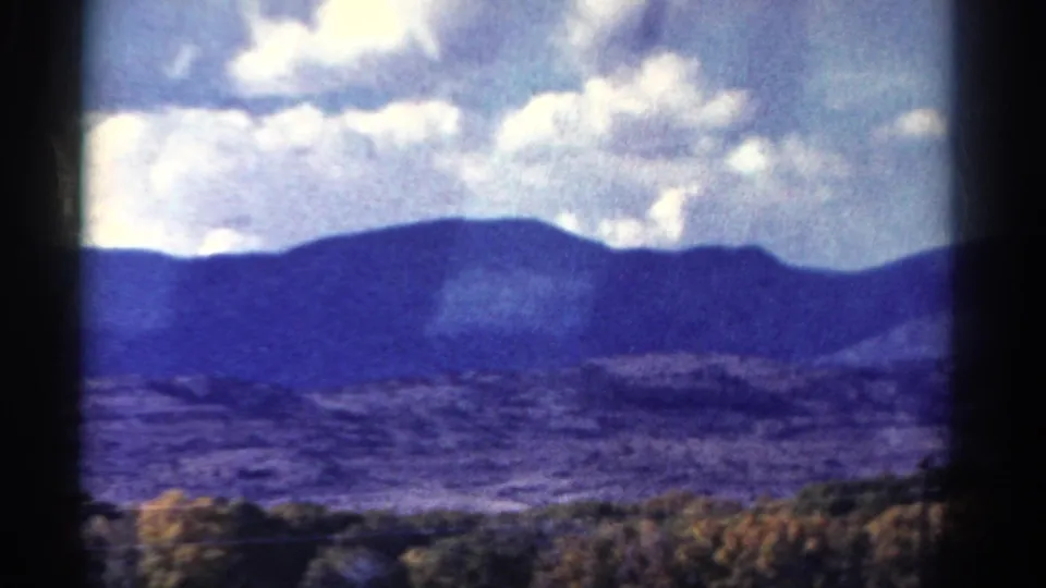 Colorado 8mm travel frame with distant mountain ridge, bright clouds, and blurred roadside trees in the foreground.