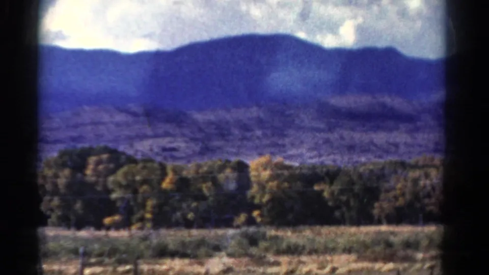 Colorado mountain ridge under broken clouds with trees and open ground in the foreground, rendered in grainy 8mm color.