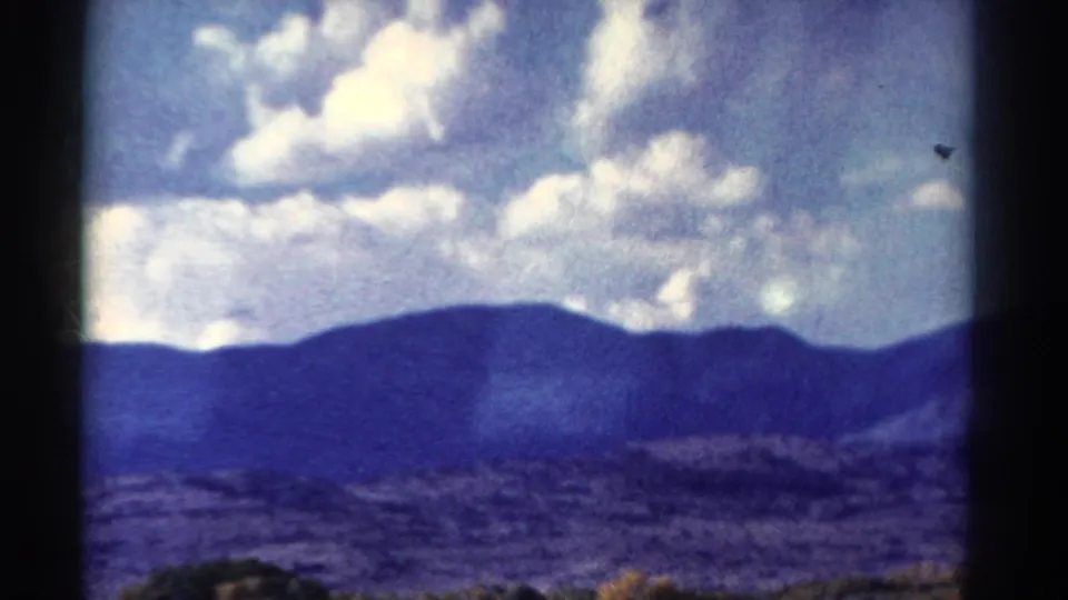 1967 Colorado home-movie frame showing mountain slopes, bright clouds, and a low tree line through a dark-edged view.