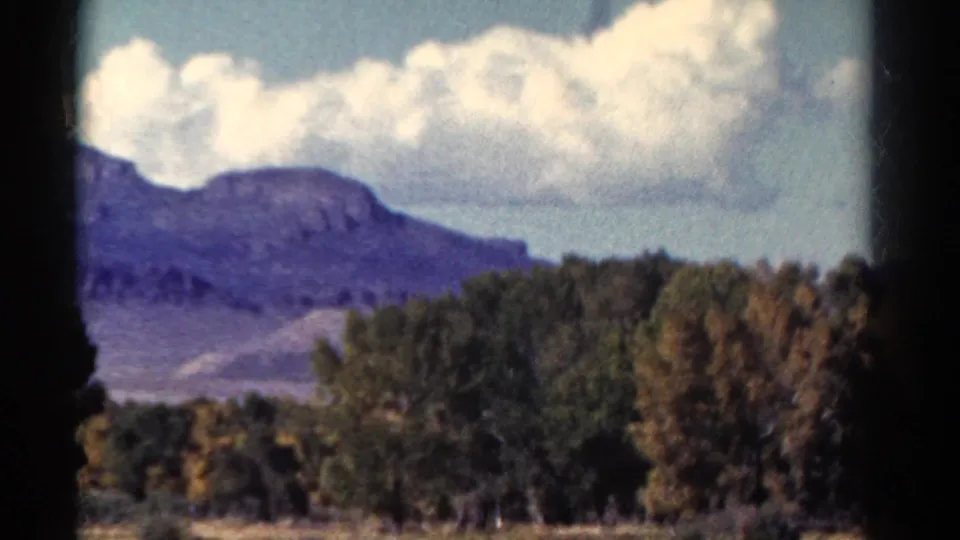 1967 Colorado home-movie frame of a mountain slope under large clouds, with trees and blurred foreground from a moving car.