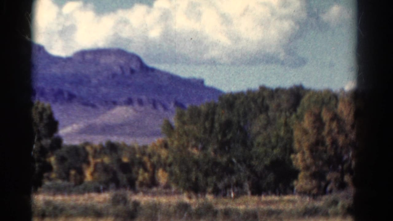 1967 Colorado 8mm travel frame showing mountains, trees, open field, clouds, and dark scan borders from a moving viewpoint.