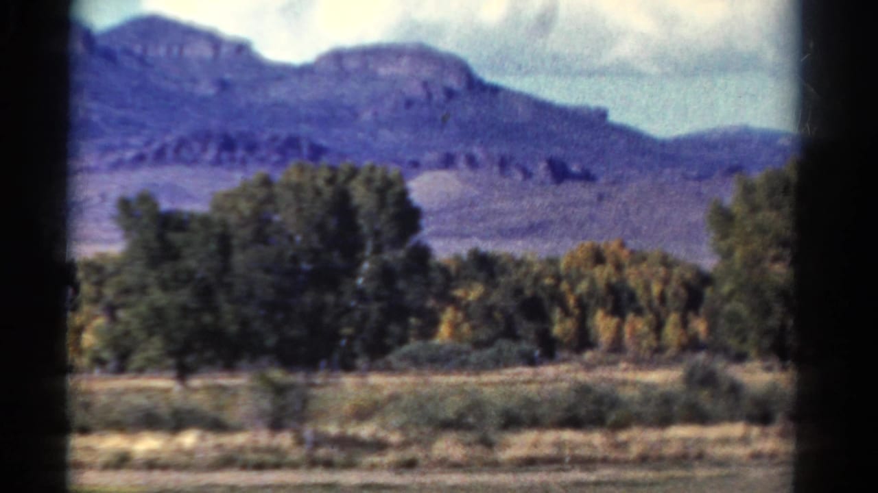 A Colorado mountain ridge rises beyond trees and open ground in this 1967 8mm passenger-window view.