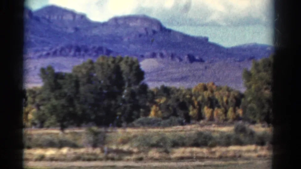 A Colorado mountain ridge rises beyond trees and open ground in this 1967 8mm passenger-window view.