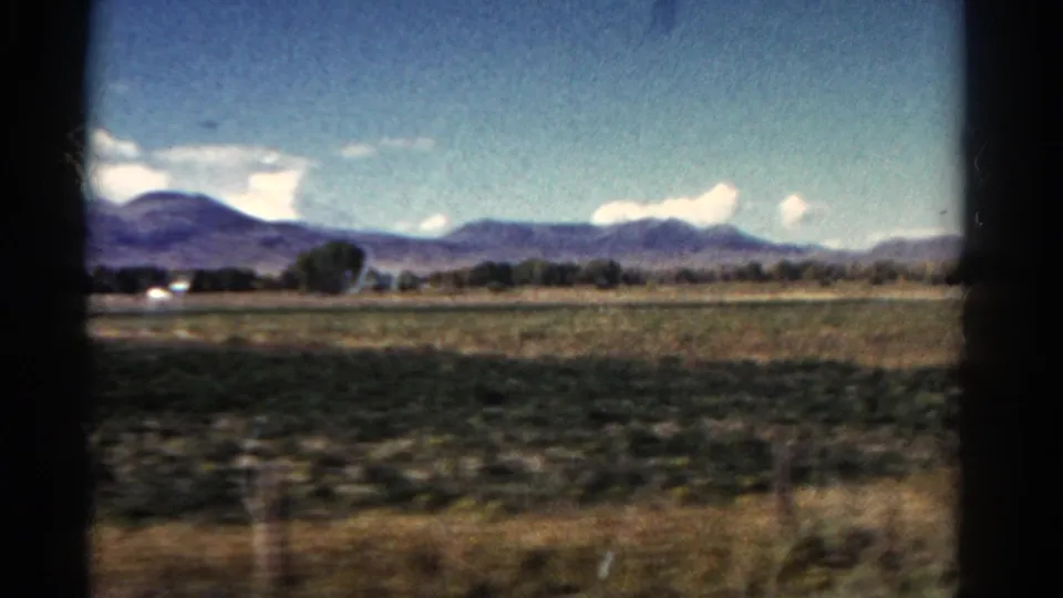 Wide 1967 Colorado countryside view with fields, low mountains, scattered trees, and foreground blur from vehicle movement.