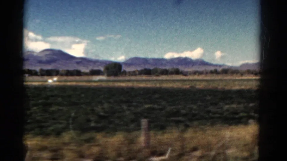 1967 Colorado 8mm view of green fields, dry roadside grass, fence posts, and distant mountains under a blue sky.