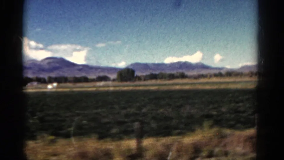 1967 Colorado roadside scene with cultivated fields, a faint road line, trees, mountains, and bright clouds in a soft 8mm frame.