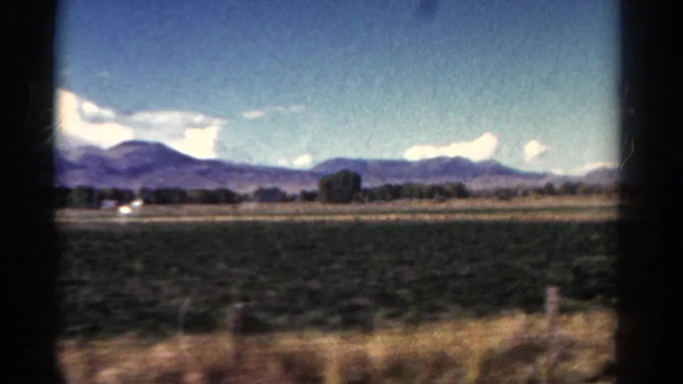 1967 Colorado 8mm frame showing a broad green field, roadside grass, fence posts, distant trees, and mountain peaks under clouds.