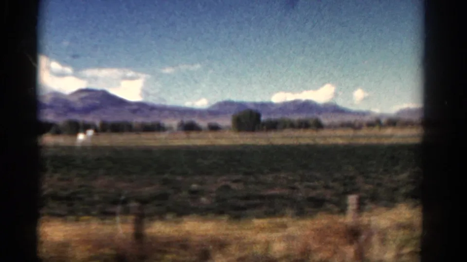 1967 Colorado home-movie frame with fields, dry shoulder grass, fence posts, a tree line, and distant mountains softened by vehicle motion.