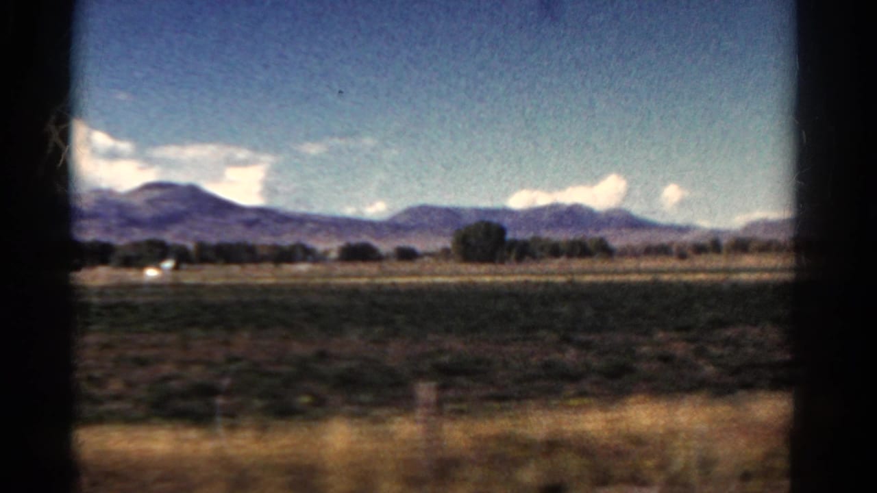1967 Colorado 8mm frame showing roadside grass, broad fields, trees, and distant mountains through a moving car window.