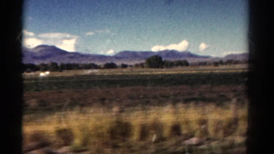 1967 Colorado travel frame with open plains, a tree line, purple-toned mountains, clouds, and dark window borders.