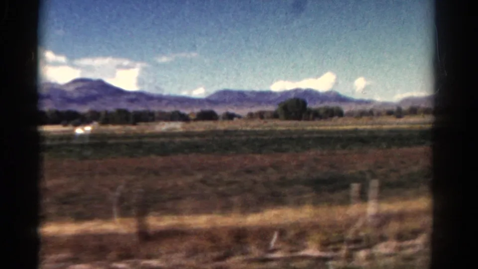 1967 Colorado 8mm view of roadside fields, distant trees and mountains, with a faint bright car-like shape at left.
