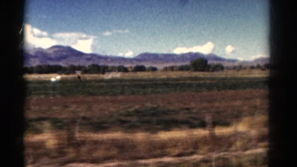 1967 Colorado home-movie frame showing fields, trees, distant mountains, clouds, and dark vertical window edges.